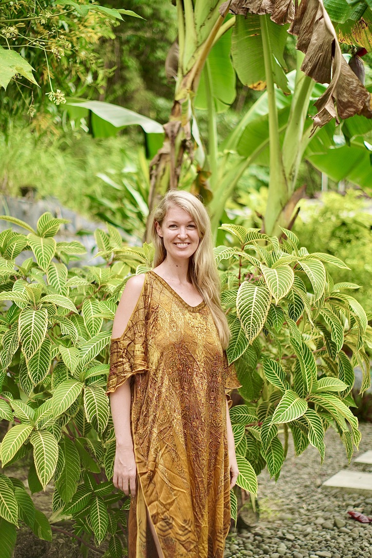 A woman with long blonde hair and an orange and brown dress stands in front of a large green plant with long, yellow-green...
