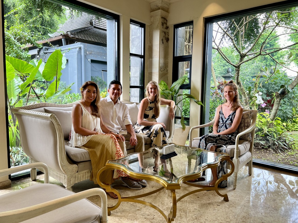 Four people sitting in a room with large windows surrounded by plants and a courtyard, three women and one man sitting in ...